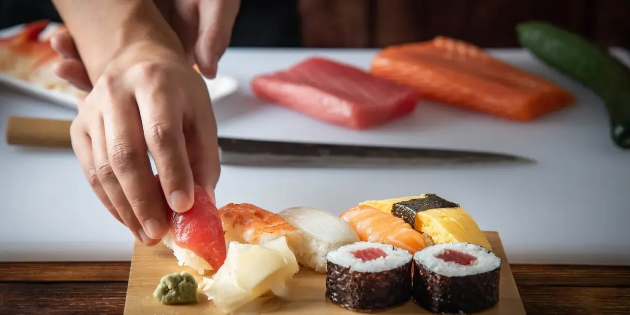 Ręce układają kawałek tuńczyka na ryżu, tworząc sushi. Na desce widać też krewetkę, kalmara, łososia i tamago. To inspiracja, jak zrobić sushi.