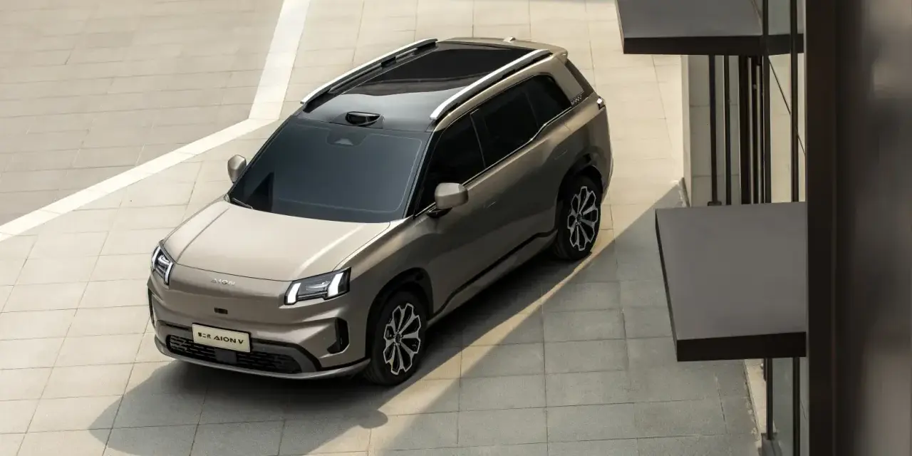 Szary elektryczny SUV GAC Aion V z czarnym dachem i relingami, zaparkowany na zewnątrz.