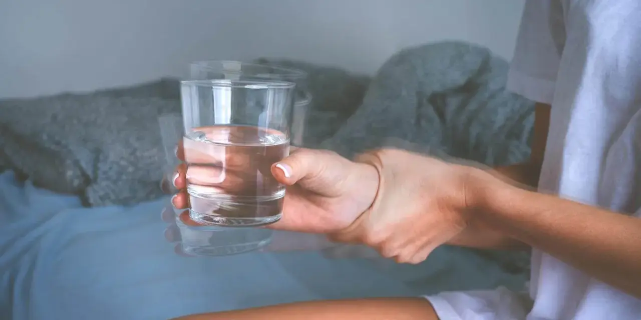 Dłoń z drżącym szkłem wody, sugerującym zwidy po alkoholu. Rozmyte kontury i niepewny chwyt.