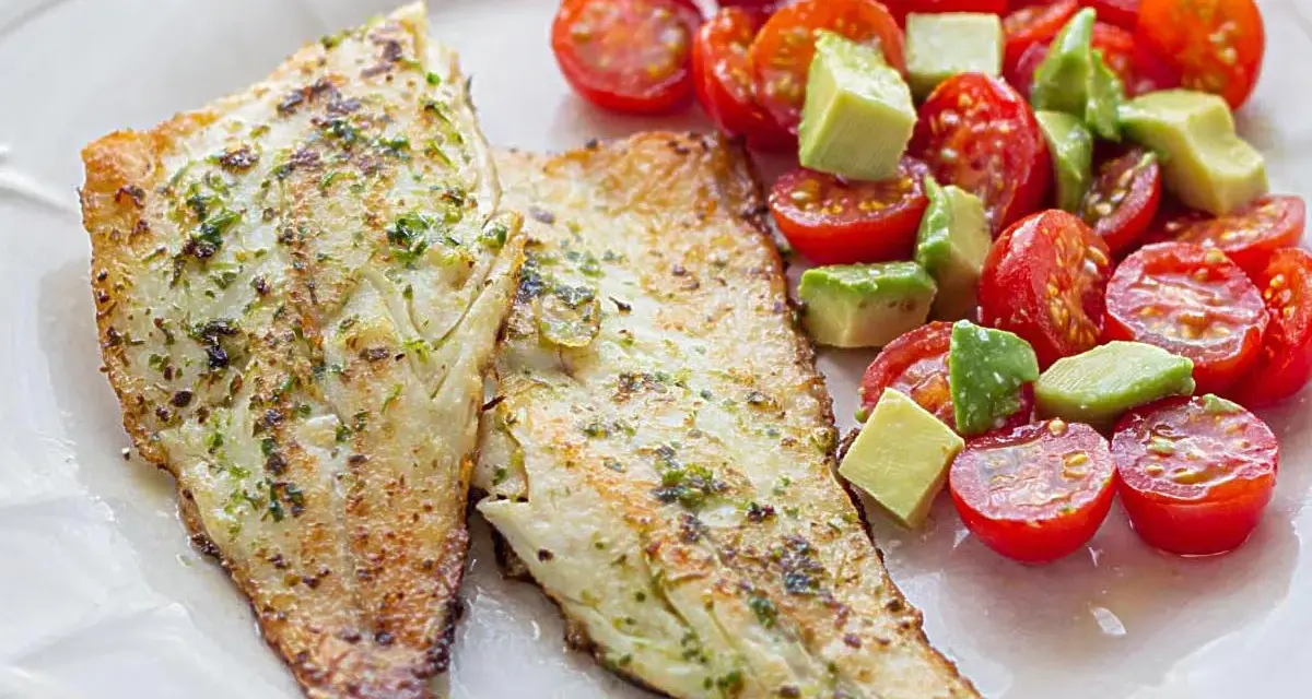 Dos filetes de lubina a la plancha con hierbas, acompañados de una ensalada fresca de tomates cherry y aguacate en cubos.