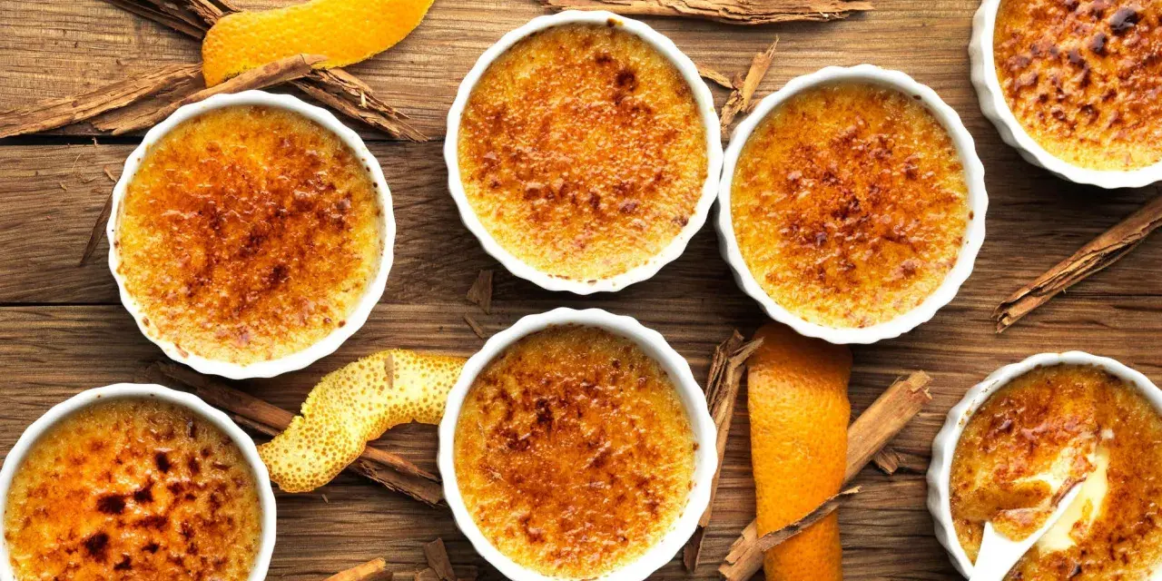 Flera krämiga crema catalana i vita skålar, toppade med karamelliserat socker. Apelsinskal och kanelstänger ger en härlig doft.