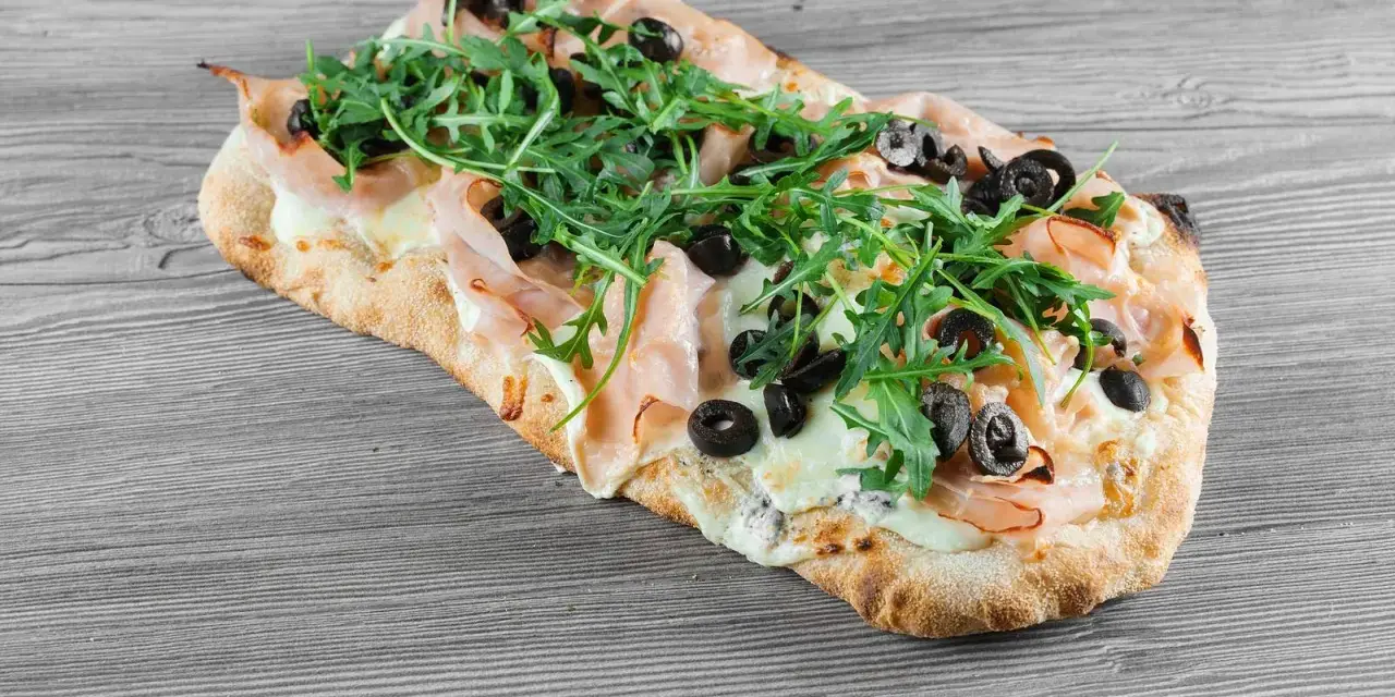 Pyszne ciasto na pizzę z szynką, mozzarellą, rukolą i czarnymi oliwkami na drewnianym tle.