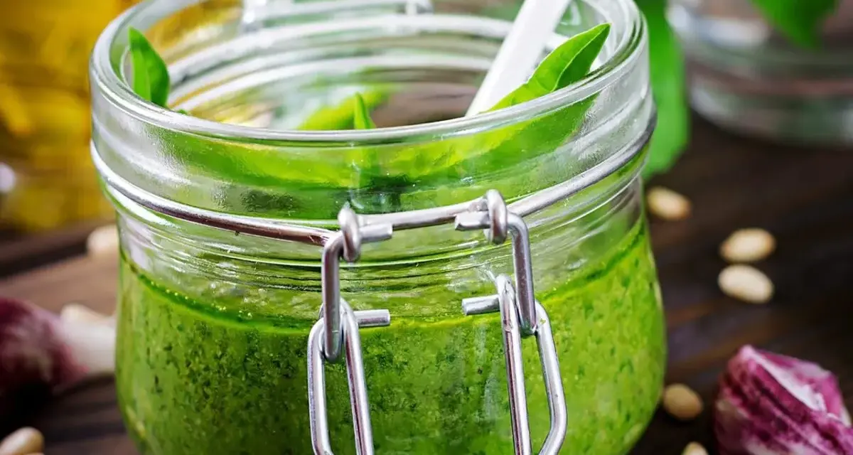 Un pot de pesto ail des ours, vert vif, avec une cuillère blanche. Des pignons de pin et de l'ail parsèment la table en bois.