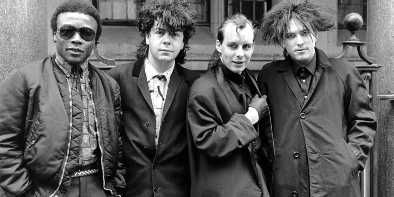 Vier Männer in dunkler Kleidung, Post-Punk-Ästhetik, stehen vor einem Gebäude.