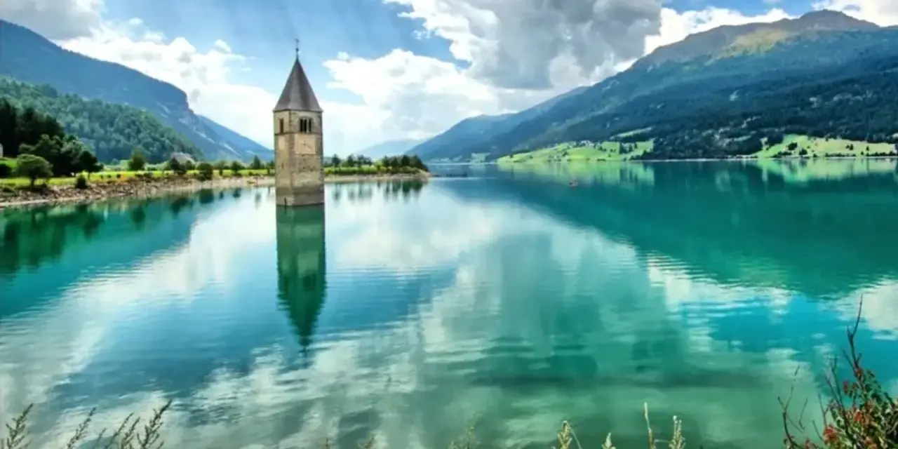 Il lago del campanile, con la sua torre sommersa, riflette montagne verdi e un cielo nuvoloso.