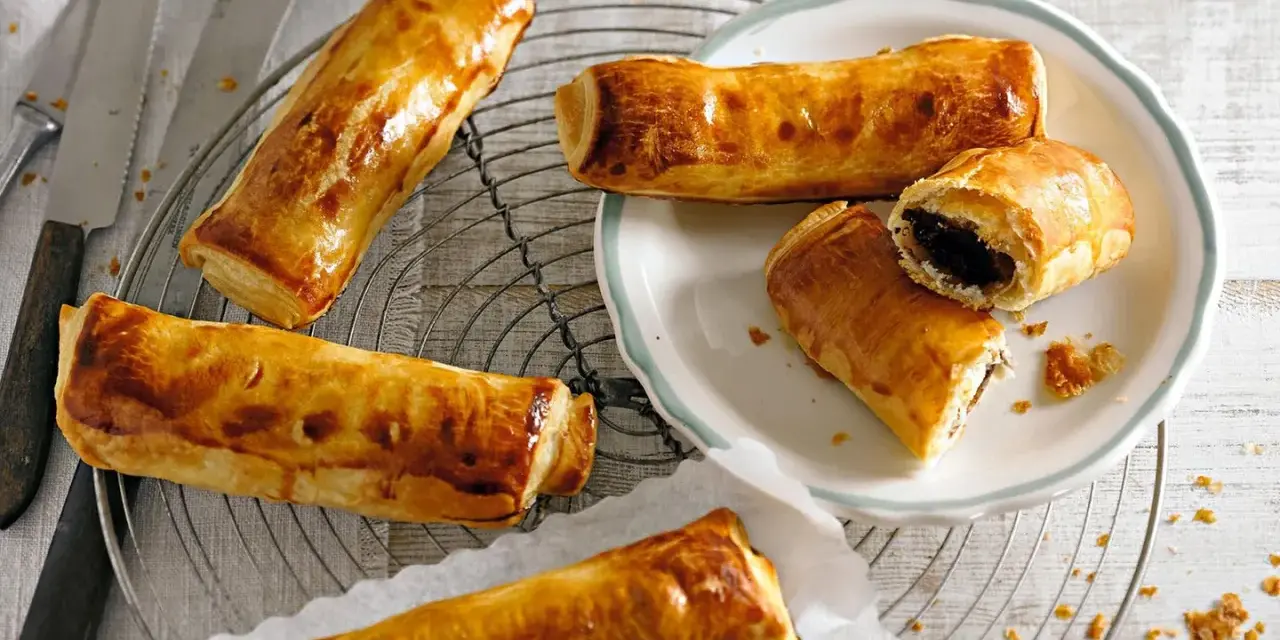 Goldbraune, knusprige Pain au Chocolat auf einem Gitter, einige auf einem Teller, einer aufgeschnitten.