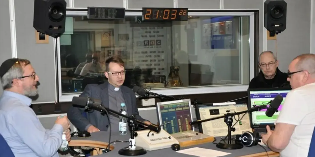 W studiu radiowym czterech mężczyzn, w tym ksiądz i osoba w jarmułce, rozmawia przy mikrofonach. W tle widać zegar i logo RDC.