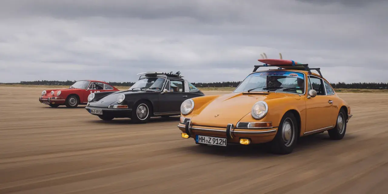 Trzy klasyczne Porsche jadące po plaży. Zdjecie w ruchu, z deskami surfingowymi na dachu jednego z nich.