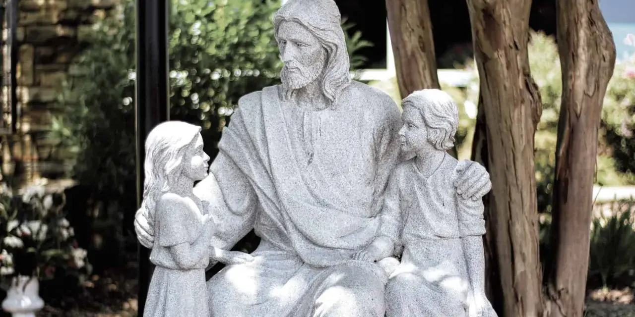 Steinfigur von Jesus, der zwei Kinder umarmt. Er wirkt wie ein liebevoller Gottvater, der seine Schützlinge beschützt.