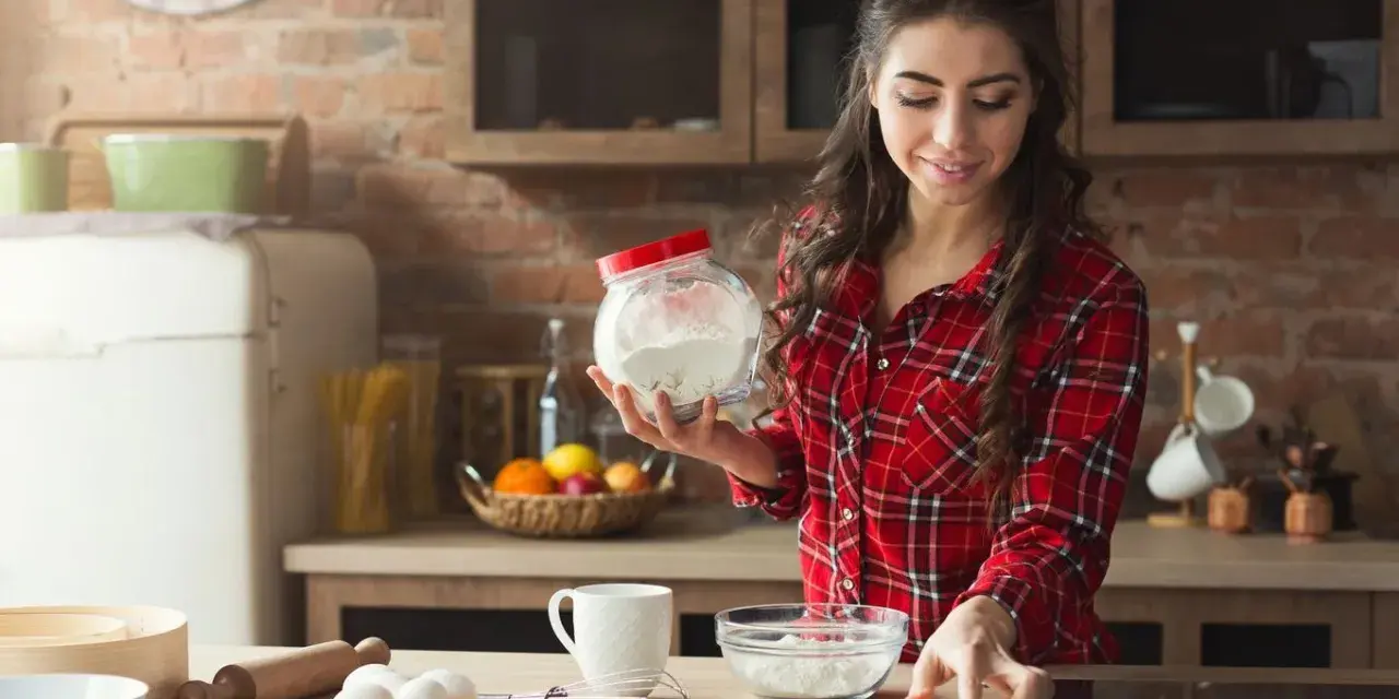 Junge Frau liest Rezept und hält Mehl bereit. Sie überlegt, ob sie Wasser statt Milch beim Backen verwenden soll.