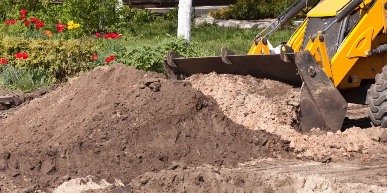 Koparka z łyżką zasypuje ziemię, przygotowując teren pod budowę. W tle kwitnące tulipany.