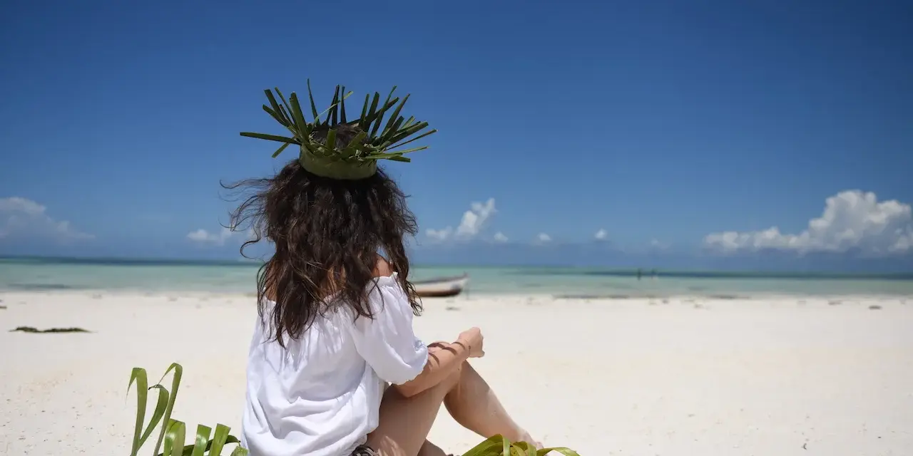 Kobieta w koronie z liści siedzi na plaży, marząc o tym, jak dolecieć na Zanzibar. Błękitne niebo i turkusowa woda zapraszają do podróży.