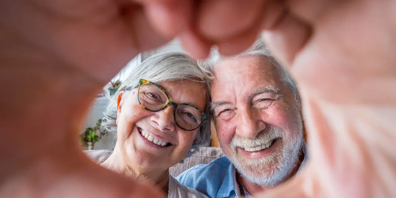 Uśmiechnięta para seniorów tworzy serce z dłoni. Ich twarze promieniują radością i miłością, która trwa przez lata.