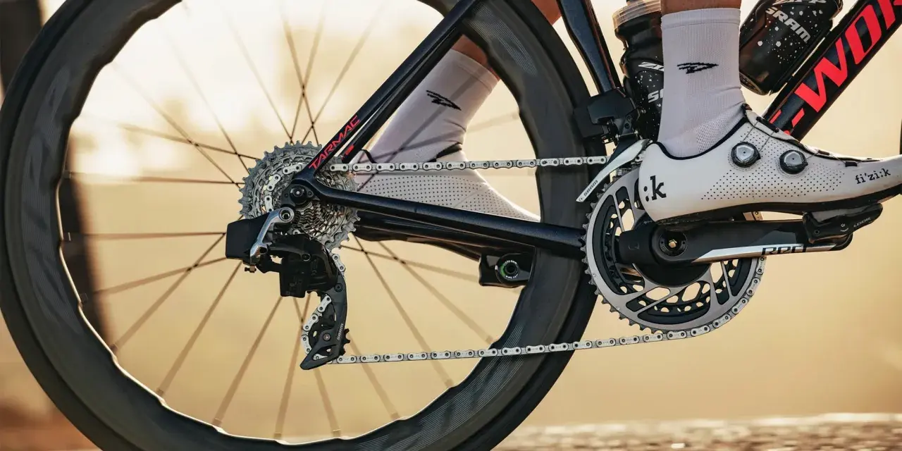 Close-up of a cyclist's shoe and drivetrain, showing the chain, gears, and crankset. The focus is on the power transfer, hinting at the cycling watts being generated.