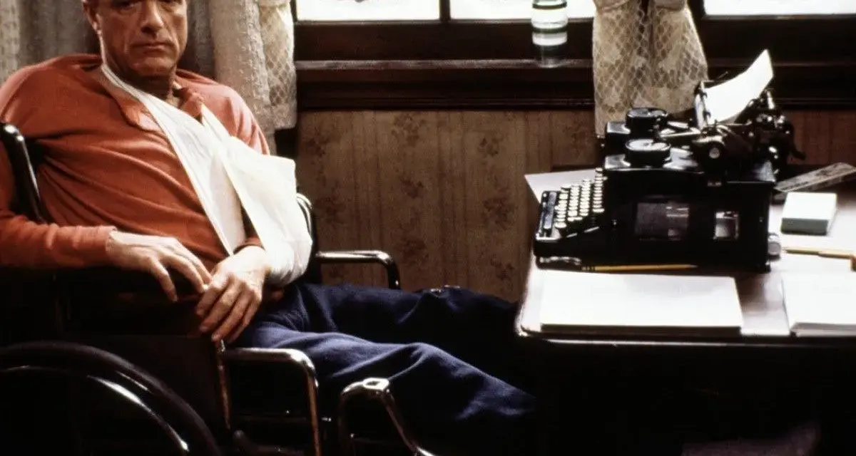 Un escritor en silla de ruedas, con un brazo en cabestrillo, mira fijamente una máquina de escribir. Escenas como esta son comunes en películas sobre escritores.