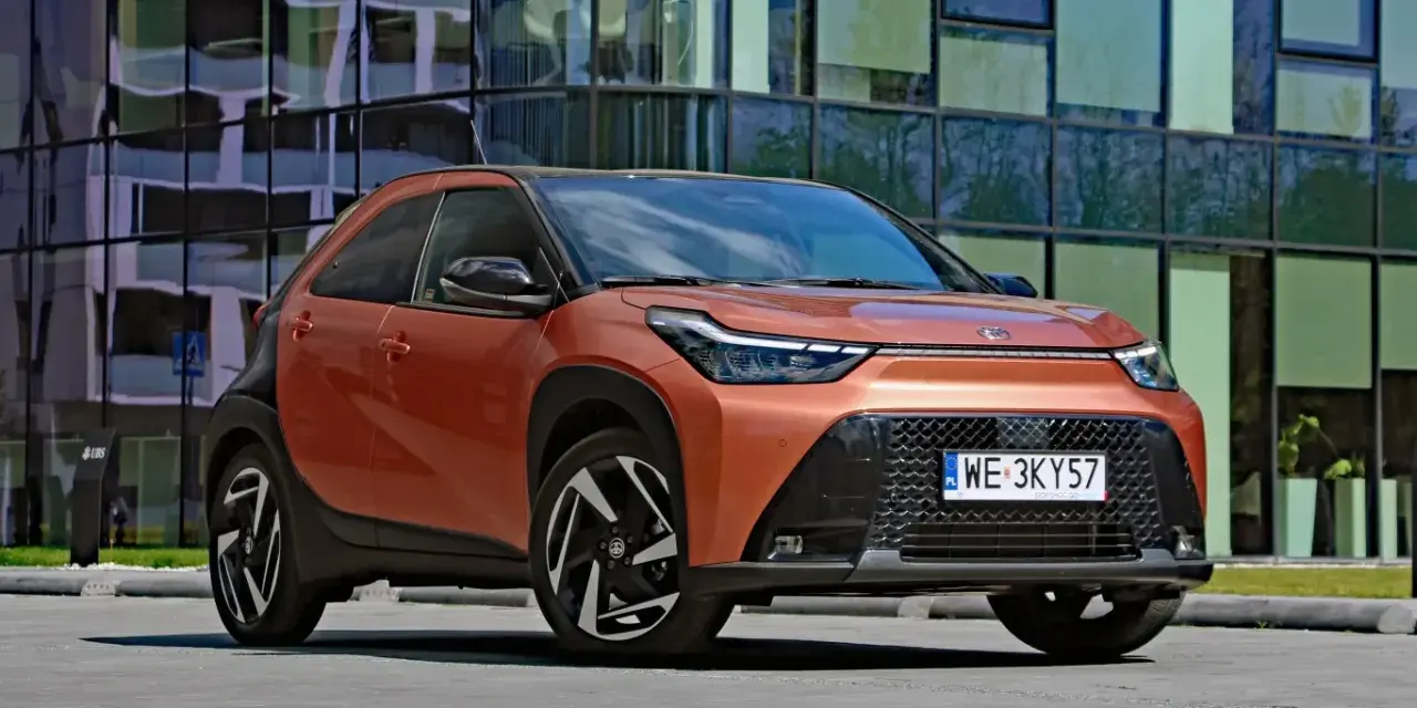 Pomarańczowy Toyota Aygo X Hybrid z czarnymi detalami stoi przed nowoczesnym budynkiem.