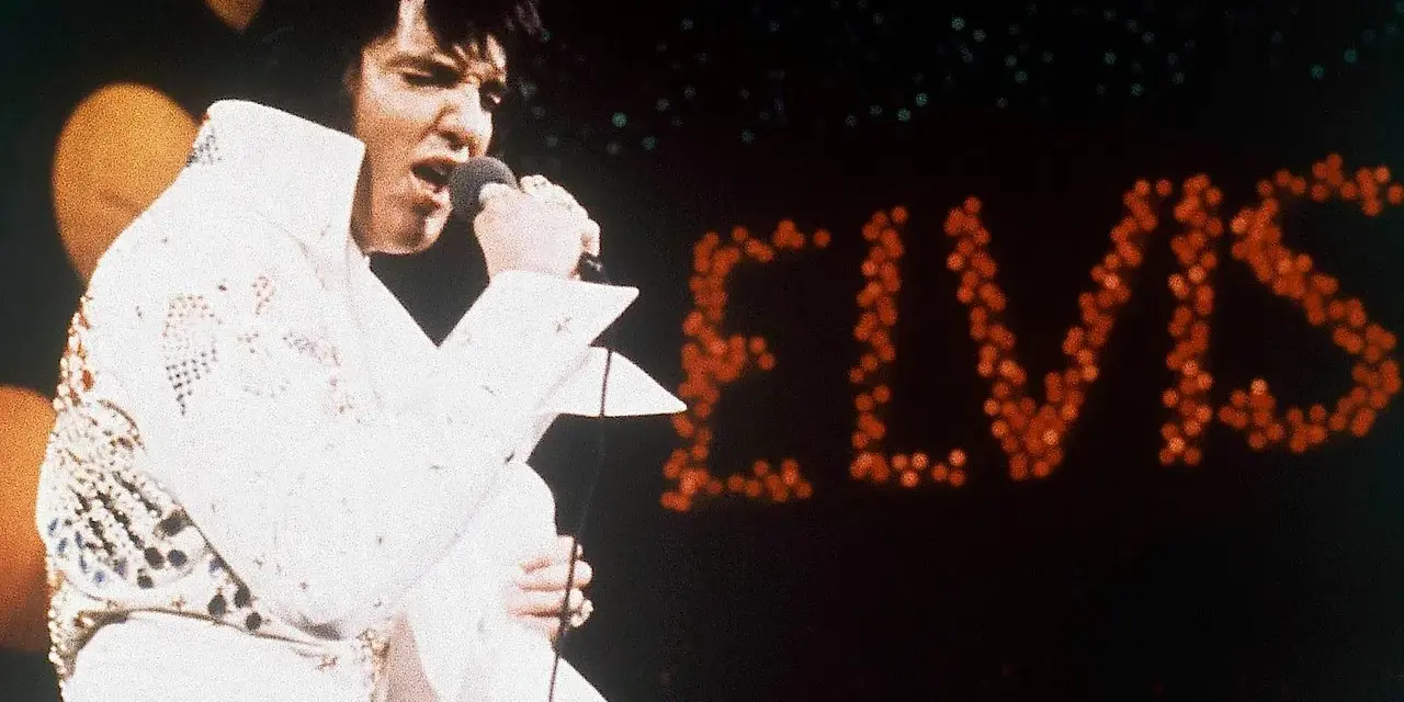 Elvis Presley singt in seinem berühmten weißen Anzug in Las Vegas, während sein Name in roten Lichtern erstrahlt.