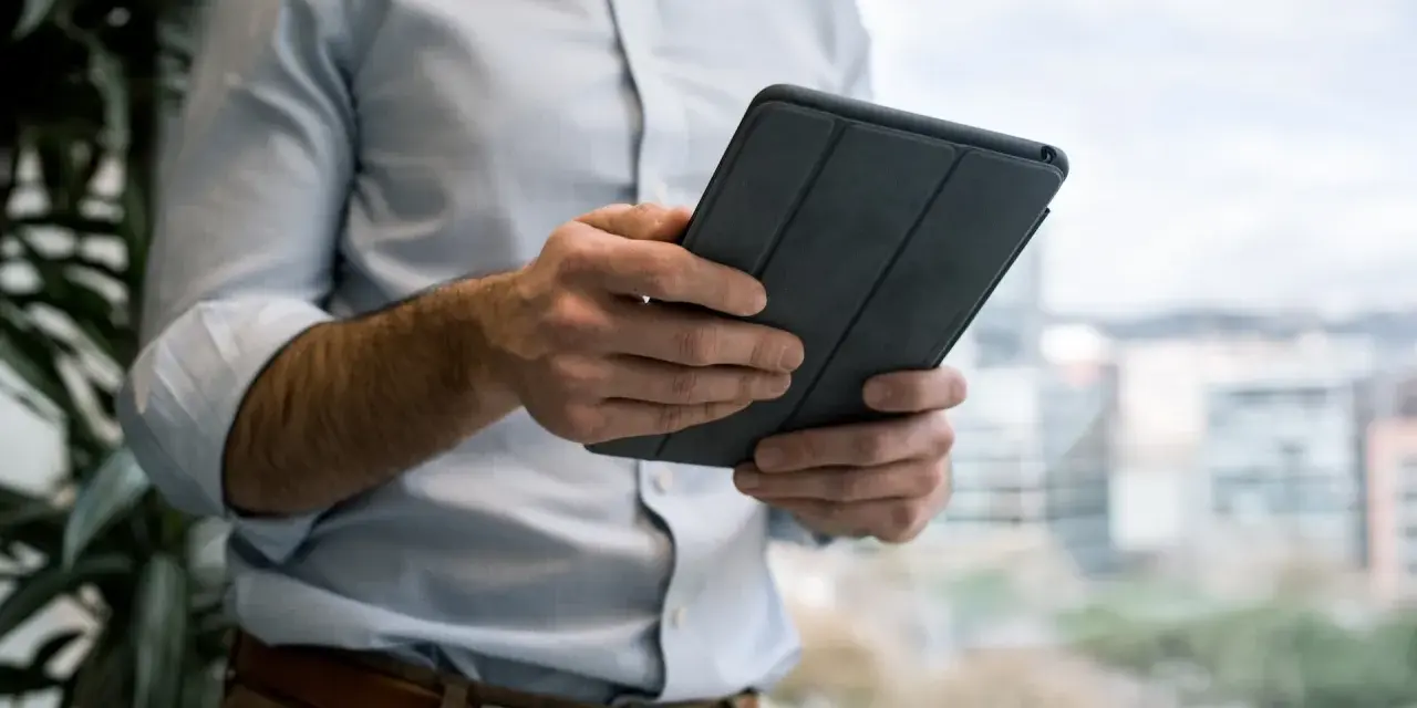 Mężczyzna w jasnej koszuli i brązowych spodniach trzyma tablet w szarej okładce. e cmr.