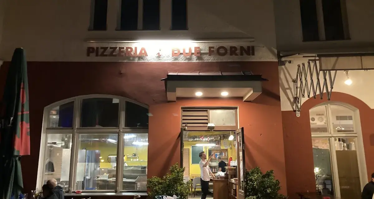Abendstimmung vor der Pizzeria "Due Forni". Ein Mann steht im Eingangsbereich, während Gäste an Tischen im Freien sitzen.