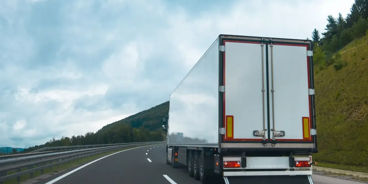 Ciężarówka na autostradzie, z tyłu widoczne białe drzwi naczepy. W tle góry i chmury. Tablice wyróżniające przepisy są częścią oznakowania pojazdu.