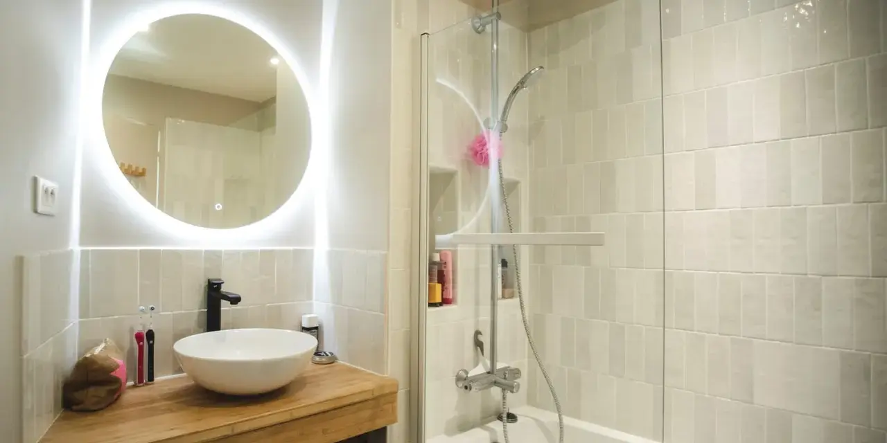 Une **salle de bain blanche** moderne avec un lavabo rond sur un meuble en bois, un miroir lumineux et une douche avec carrelage clair.