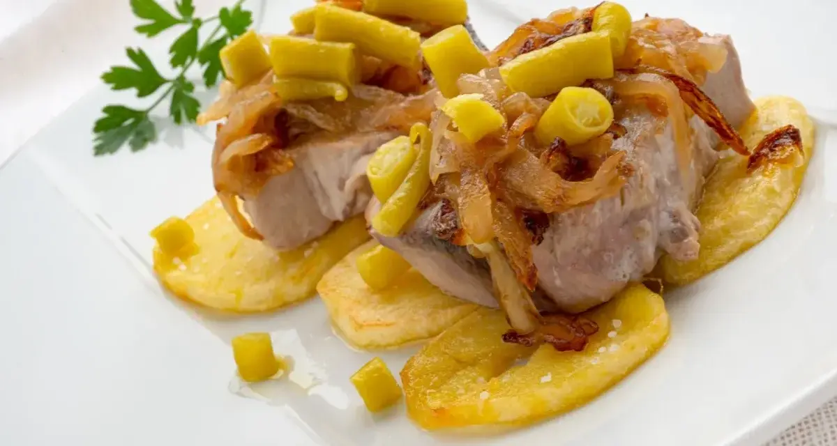 Bonito al horno sobre patatas, con cebolla caramelizada y pimientos verdes. Un plato delicioso y bien presentado.