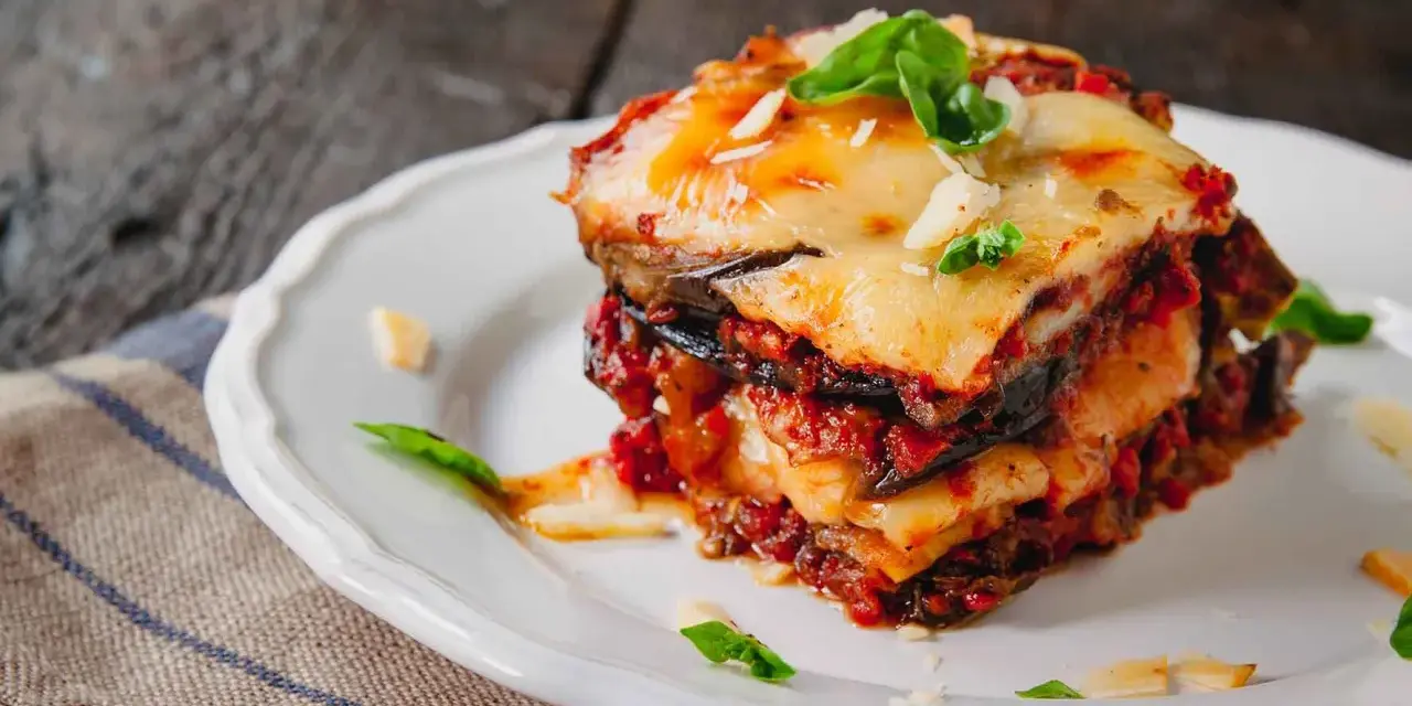 Una fetta di parmigiana di melanzane al forno, stratificata con melanzane, sugo e formaggio filante, guarnita con basilico fresco.