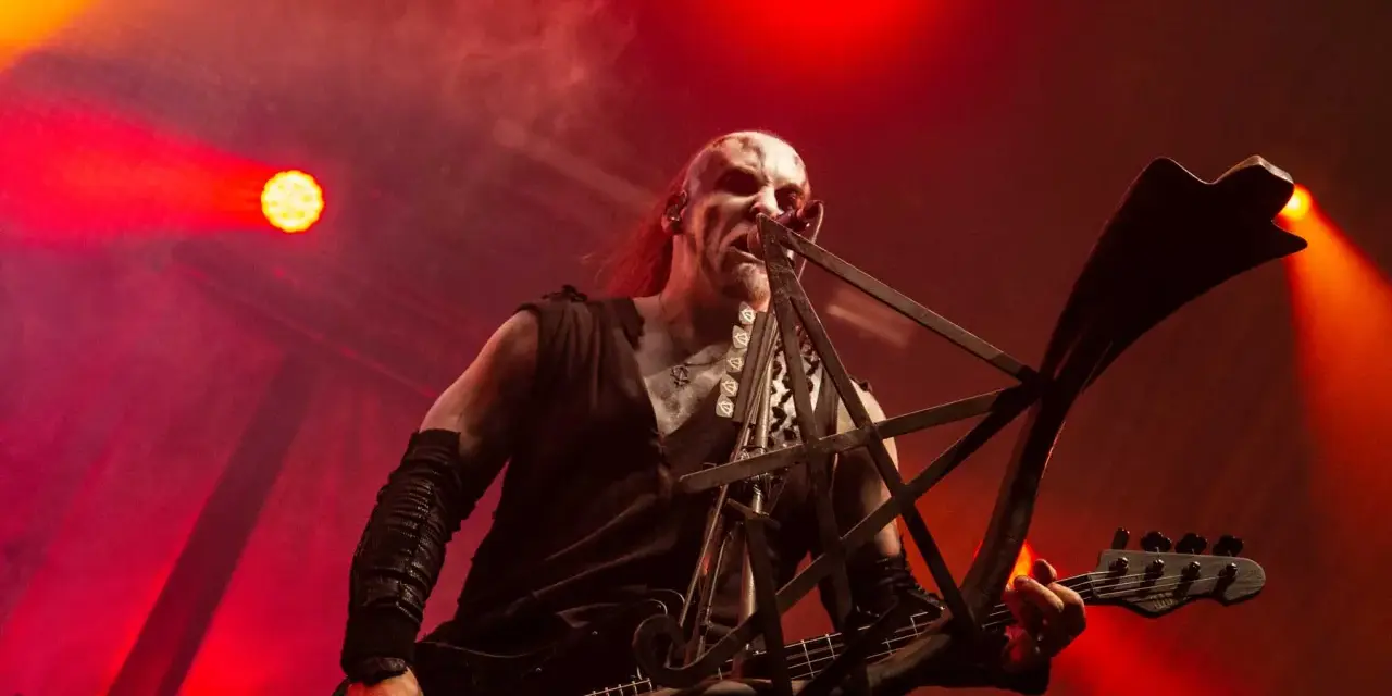 Un musicien au visage peint, jouant de la basse sur scène, baigné de lumières rouges. Sa posture exprime une vie d'agonie.