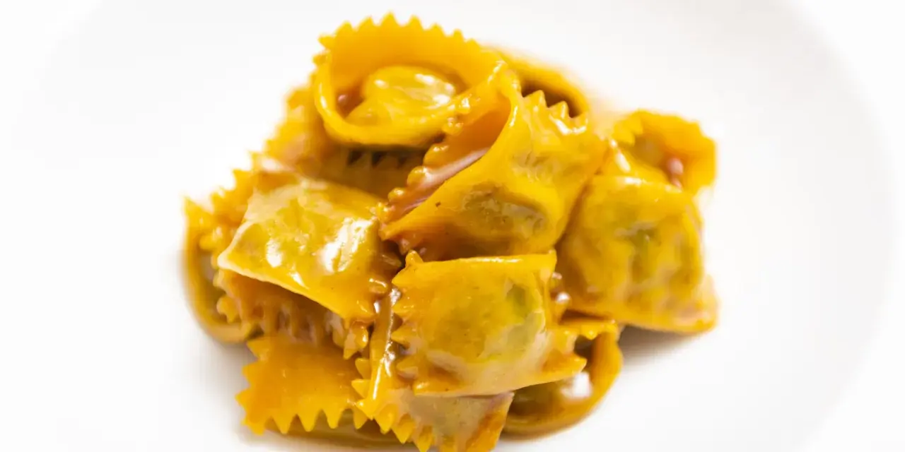 Un mucchietto di ravioli del plin, dorati e conditi con un sugo cremoso, su un piatto bianco.