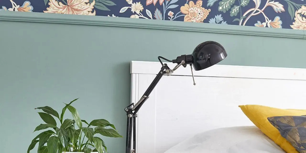 Zielona lamperia pod kwiecistną tapetą, obok łóżka stoi lampka i doniczka z rośliną.