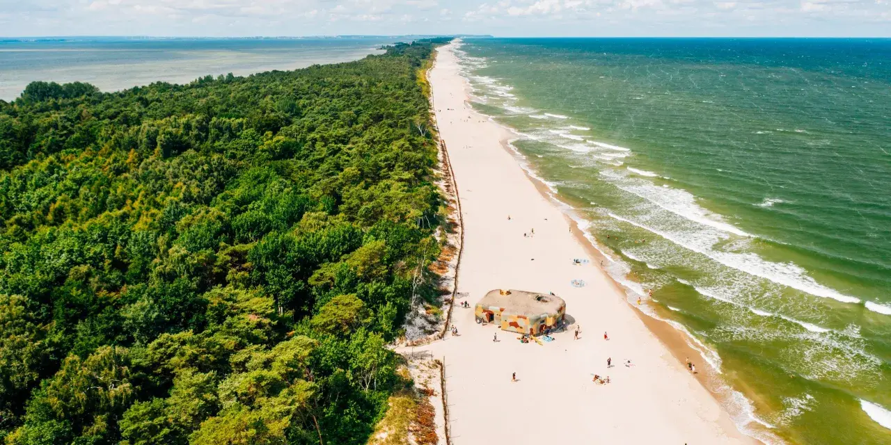 Majówka nad morzem: zielony las, piaszczysta plaża z kilkoma osobami i bunkrem w kamuflażu, szmaragdowe fale Bałtyku.