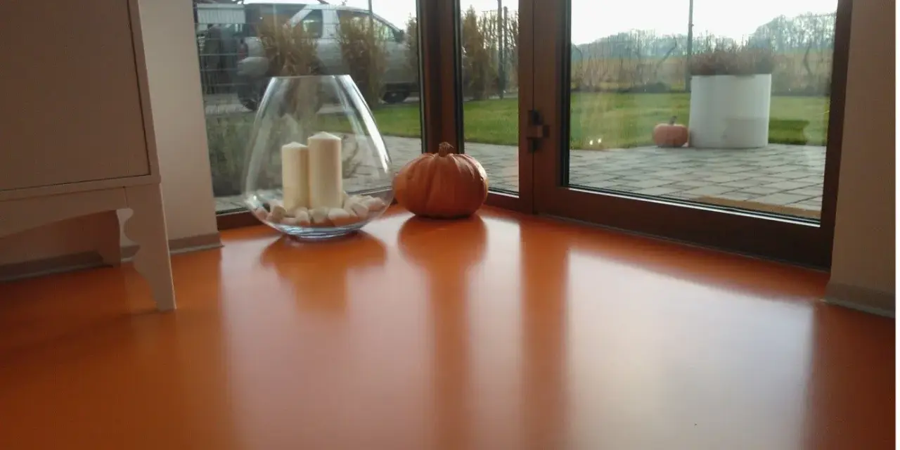 Pomarańczowa podłoga z żywicy, z widocznym blaskiem i odbiciami. Wazon ze świecami i dynia dodają jesiennego klimatu.