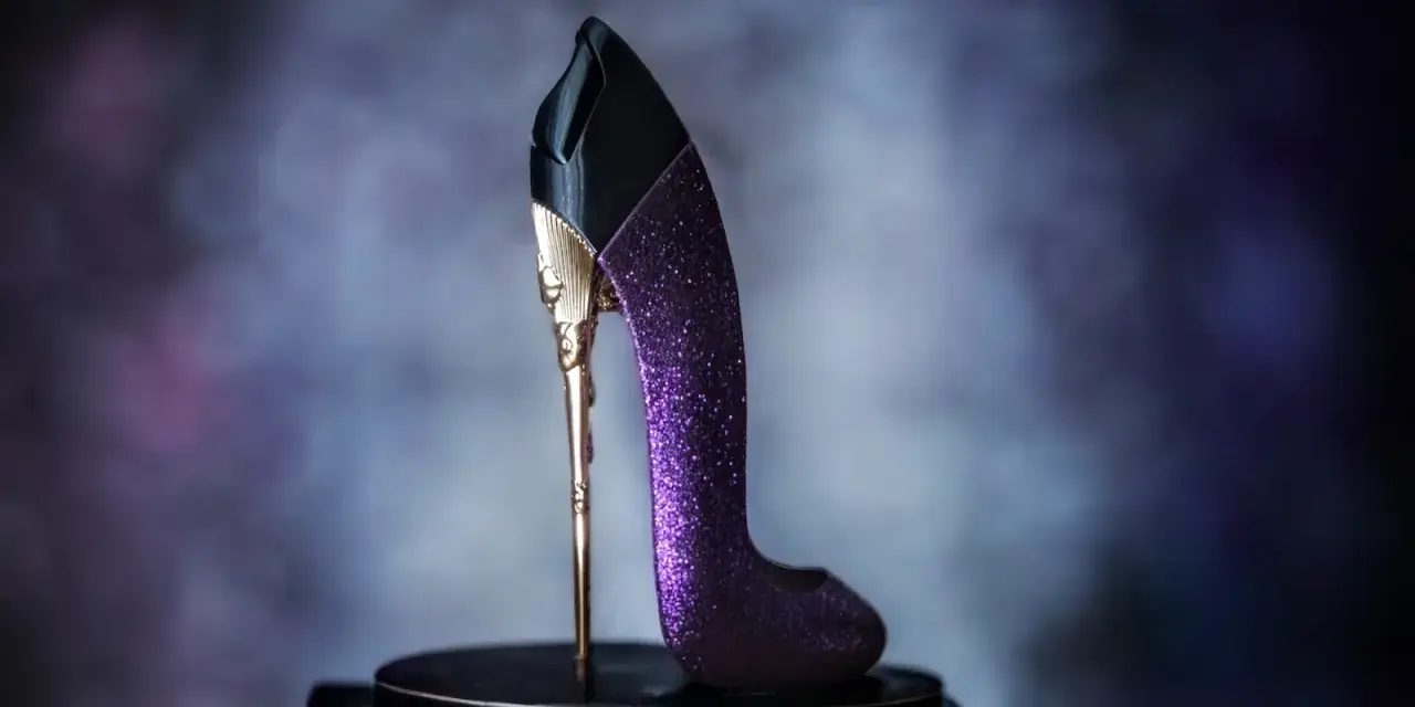 Elegancki perfumy bucik w kolorze fioletowym, ozdobiony złotymi detalami i brokatem, stoi na czarnym postumencie.