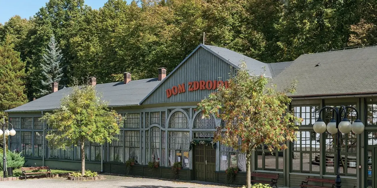 Długopole Zdrój atrakcje: zabytkowy Dom Zdrojowy z czerwonym napisem, otoczony zielenią i drzewami.