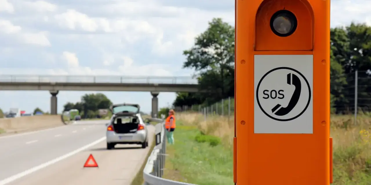 Ihr Fahrzeug ist auf der Autobahn liegen geblieben. Eine Frau in Warnweste steht neben dem offenen Kofferraum, ein Warndreieck liegt auf der Fahrbahn. Eine Notrufsäule steht im Vordergrund.