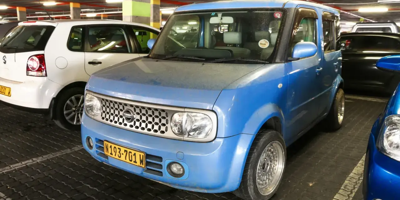 Niebieski Nissan Cube z charakterystycznym grillem i tablicą rejestracyjną N193-701 W, zaparkowany obok białego Volkswagena.