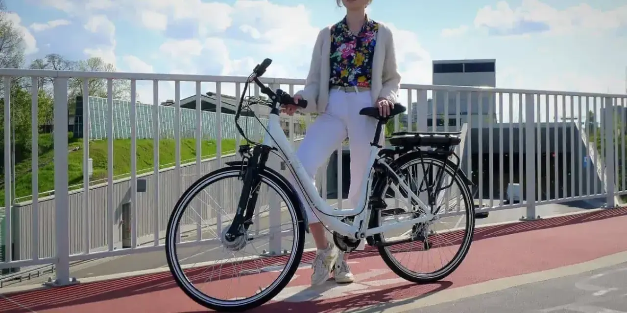 Kobieta w białych spodniach i kolorowej koszuli stoi obok białego roweru elektrycznego. Czy na rower elektryczny trzeba mieć prawo jazdy?