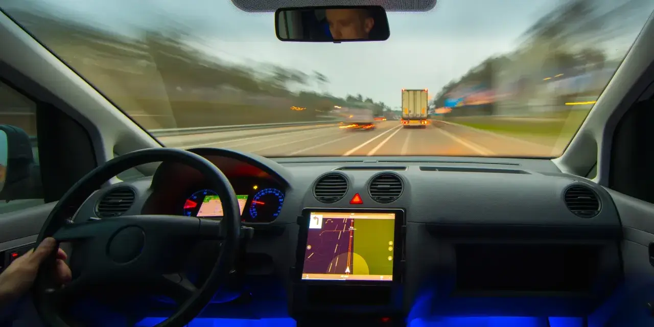 Widok z kabiny samochodu jadącego autostradą. Na desce rozdzielczej tablet z nawigacją, a obok niego system rozpoznawania znaków drogowych.