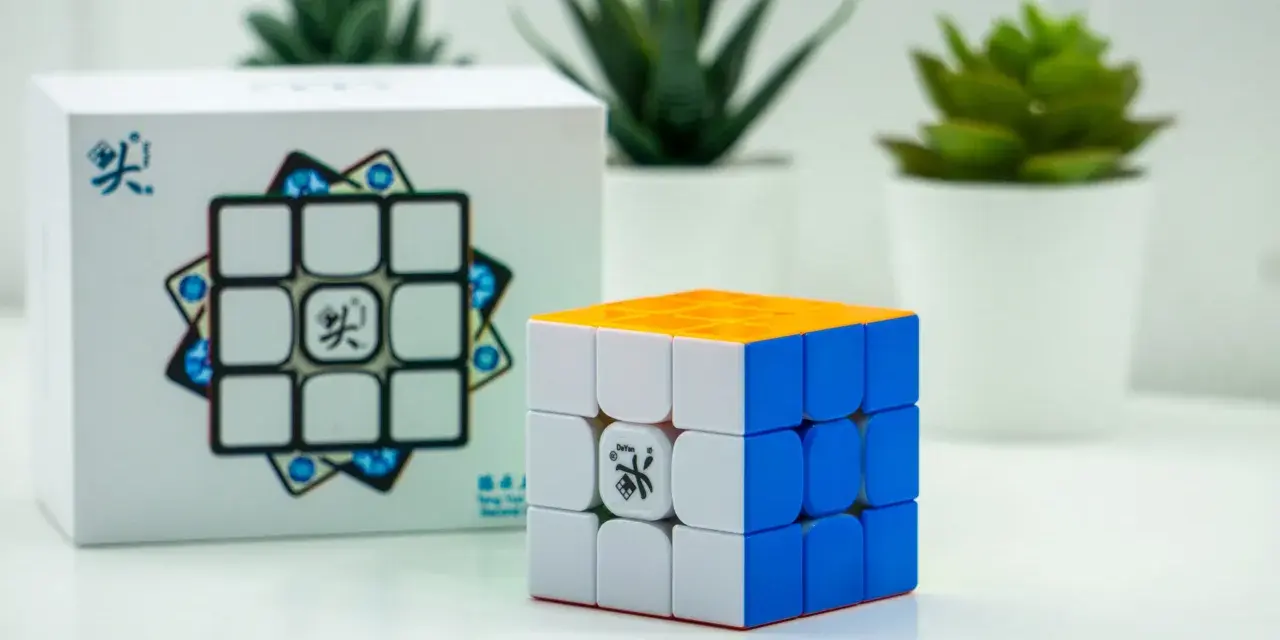 En speed cube 3x3 med orange, vit och blå sida står bredvid sin förpackning. Tre gröna växter i vita krukor syns i bakgrunden.