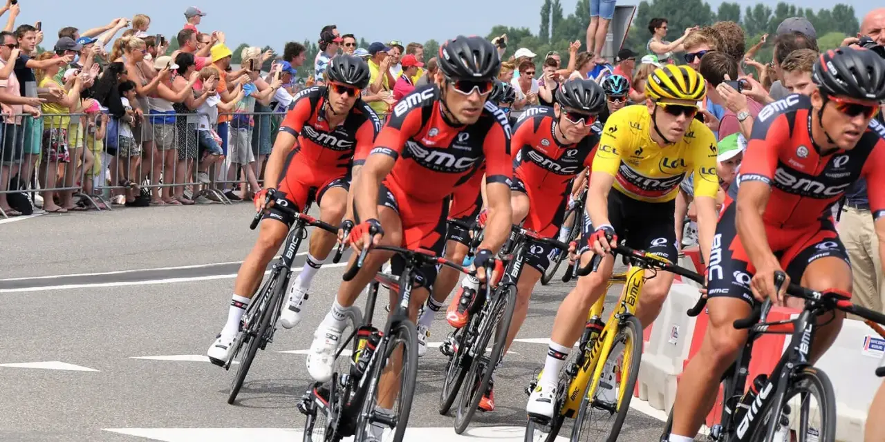 Cyclists in red BMC jerseys race, with one in yellow, as spectators cheer. A perfect scene to watch Tour de France live!