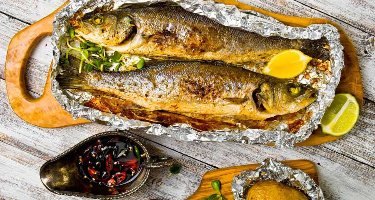 Due branzini cotti al cartoccio, dorati e succulenti, serviti con spicchi di limone, patata al cartoccio e salsa piccante.