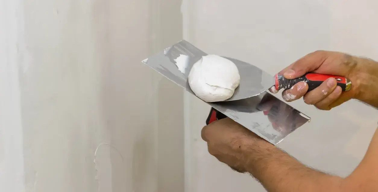 Mani che usano spatole per applicare stucco su un muro. Lo stucco serve a livellare e riparare le imperfezioni.