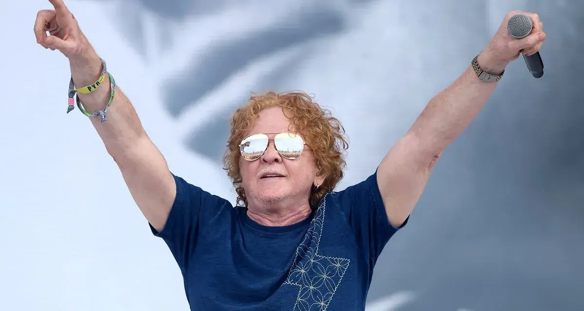Wokalista Simply Red z uniesionymi rękami, trzymający mikrofon. Ma charakterystyczne rude włosy i okulary przeciwsłoneczne.