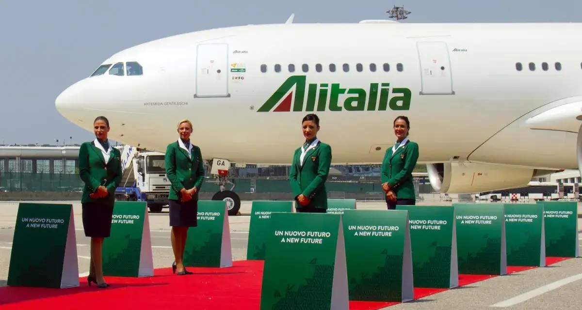 Cztery stewardessy w zielonych uniformach stoją na czerwonym dywanie przed samolotem Alitalia.