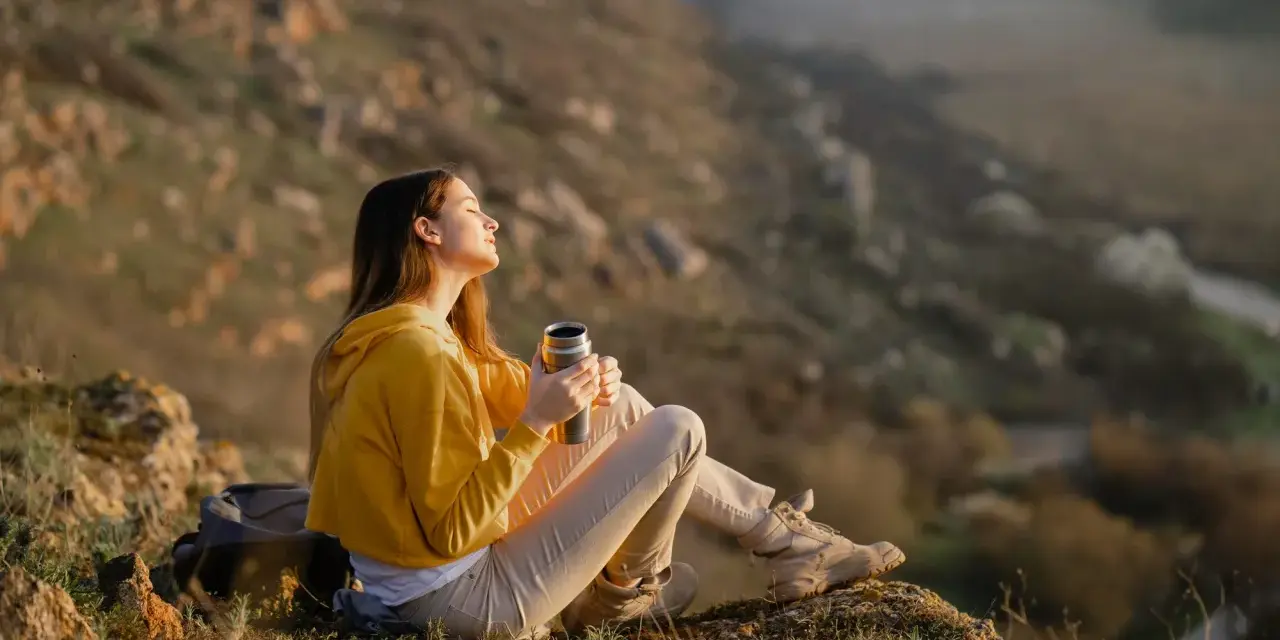 A young woman enjoys the art of doing nothing, holding a thermos and basking in the sun on a rocky hillside.