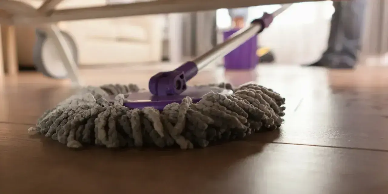 Mop pod łóżkiem, gotowy do czyszczenia paneli. W tle widać wiadro i nogi osoby sprzątającej.