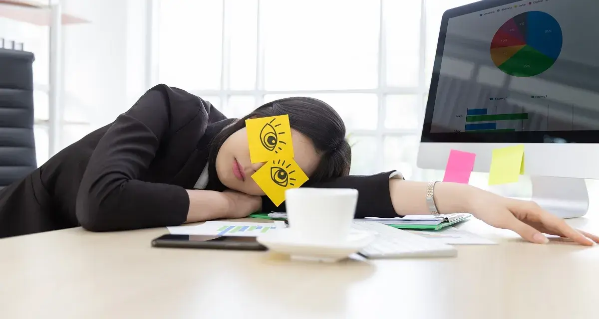 En kvinna sover vid sitt skrivbord med post-it-lappar som föreställer ögon på ögonlocken. Det är uppenbart att hon tycker det är tråkigt på jobbet.