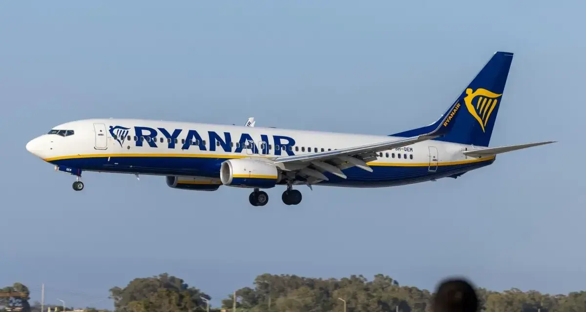 Samolot Ryanair lądujący na tle nieba i drzew, z widoczną sylwetką osoby w pierwszym planie.