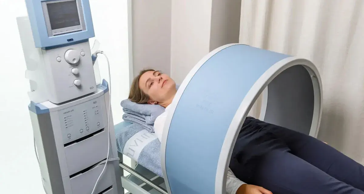 Kobieta leży na łóżku rehabilitacyjnym, poddając się zabiegowi z wykorzystaniem urządzenia generującego pole magnetyczne, które obejmuje jej kolano.
