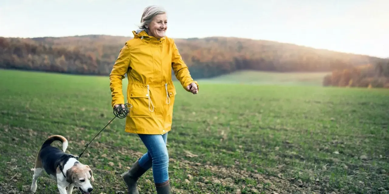 Glad kvinna i gul jacka njuter av en promenad med sin hund i ett fält. En härlig promenad som bränner kalorier.